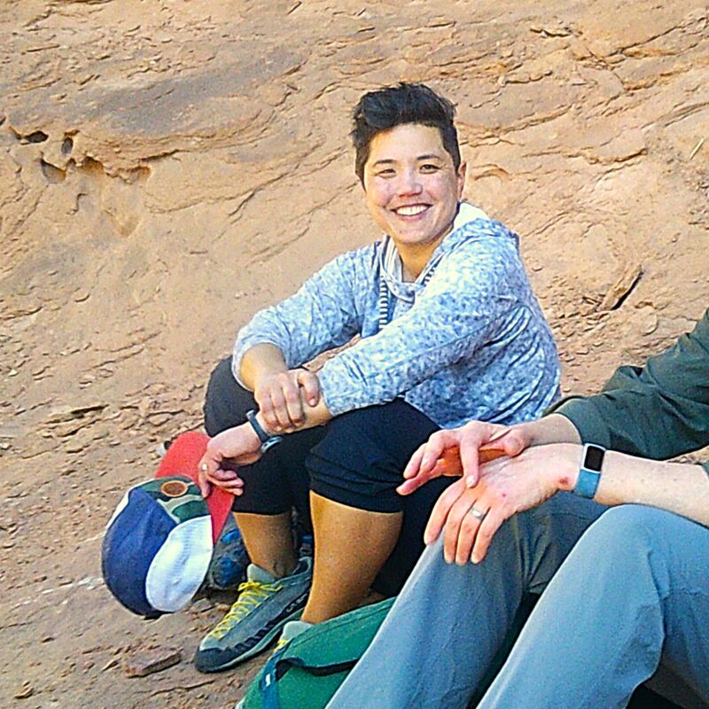 A person with short dark hair is sitting on the ground, smiling at the camera. They are wearing a light blue patterned hoodie, black pants, and climbing shoes. Next to them is a colorful hat and another person wearing blue pants and a watch. The background appears to be a rocky, outdoor setting.